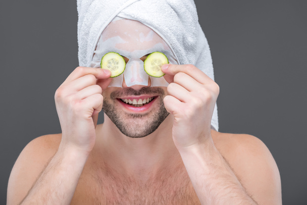 Улыбающийся бородатый мужчина с коллагеновой маской и огуречными ломтиками на глазах, изолированный на сером
 - Фото, изображение