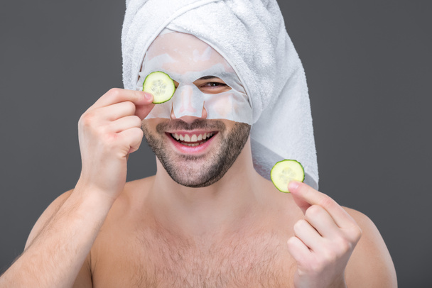 vrolijke bebaarde man met collageen masker en plakjes komkommer, geïsoleerd op grijs, huid zorg concept - Foto, afbeelding