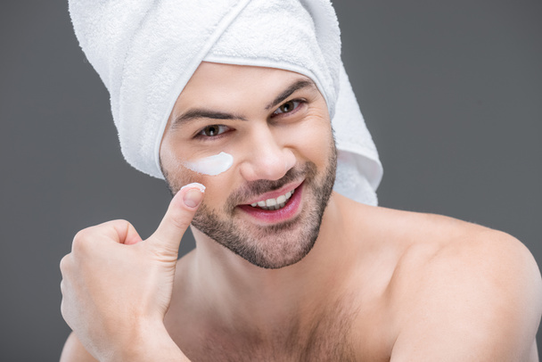 cheerful beardman in towel applying face cream, isolated on grey, skin care concept - Foto, Imagem