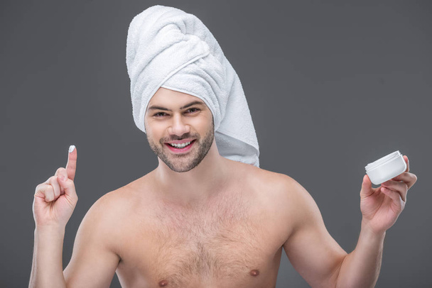 cheerful beardman in towel holding face cream, isolated on grey - Photo, Image
