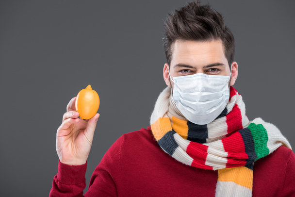 hombre enfermo en bufanda caliente y máscara médica sosteniendo limón, aislado en gris
 - Foto, Imagen