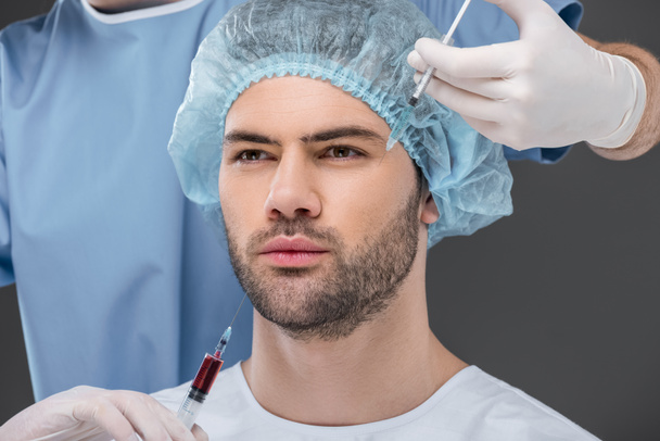 бородатый красивый мужчина в медицинском колпаке делает инъекции красоты, изолированные на сером
 - Фото, изображение