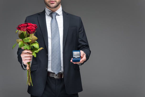 cropped view of man holding bouquet of red roses and proposal ring, isolated on grey - Fotografie, Obrázek