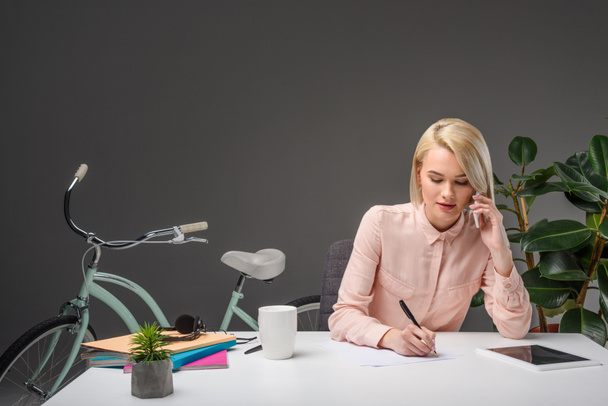retrato de la mujer de negocios hablando en el teléfono inteligente mientras toma notas en el lugar de trabajo
 - Foto, Imagen