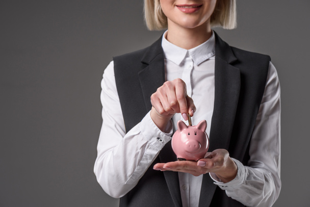 cropped shot of businesswoman putting coin into piggy bank isolated on grey - Photo, Image