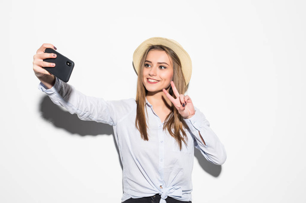 Retrato de uma jovem mulher atraente com chapéu fazendo foto selfie no smartphone com sinal de vitória isolado no fundo branco
 - Foto, Imagem