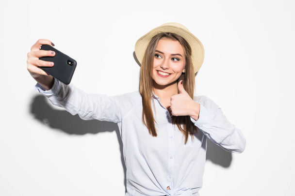 Porträt einer jungen attraktiven Frau mit Hut macht Selfie-Foto auf Smartphone mit Daumen nach oben isoliert auf weißem Hintergrund - Foto, Bild