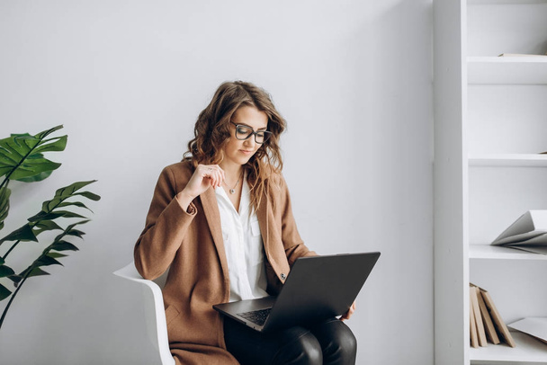 Дивовижна бізнес-леді сидить на стільці з ноутбуком у своєму офісі з яскравим інтер'єром
 - Фото, зображення