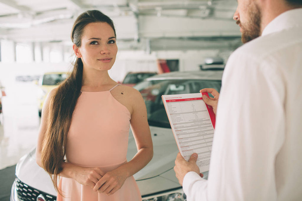 vendedor de coches amigable hablando con una mujer joven y mostrando un coche nuevo dentro de la sala de exposición Firma del contrato
. - Foto, imagen