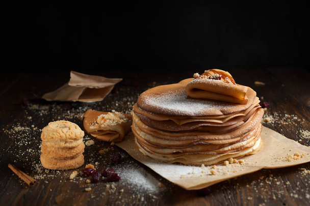 stapel pannenkoeken, gevouwen pannenkoek, besprenkeld met bessen, cookies, poeder suiker en karamel op perkament op een donkere houten tafel - Foto, afbeelding