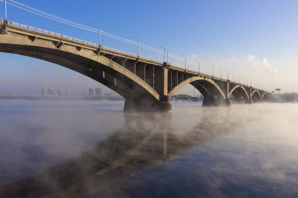 Αστικό τοπίο με αψίδα κοινόχρηστη γέφυρα πάνω από τον ποταμό Yenisei σε Κρασνογιάρσκ χειμώνα καταπληκτική βραδιά. Η γέφυρα αυτή αντικατοπτρίζεται στην καθρέφτης-όπως επιφάνεια του νερού - Φωτογραφία, εικόνα