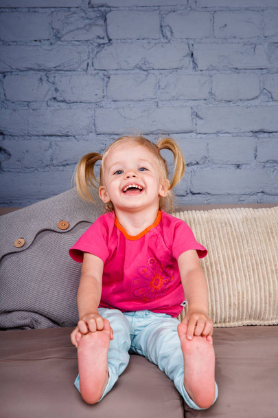 mała dziewczynka śmieszne zanurza się na kanapie w domu z rękami za jej obcasy. Śmieje się, pokazując jego język i zęby. Ubrana w jasne stylowe ubrania - Zdjęcie, obraz