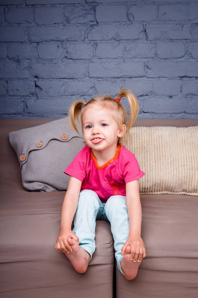 the little funny girl indulges on the sofa at home with her hands behind her heels. He laughs showing his tongue and teeth. Dressed in bright stylish clothes - Фото, изображение