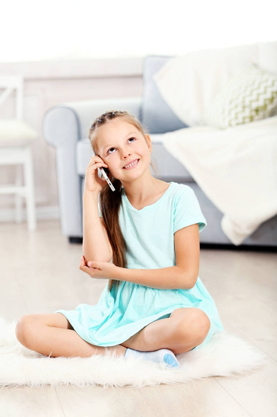Little cute girl with mobile phone sitting on carpet, on home interior background - Fotoğraf, Görsel