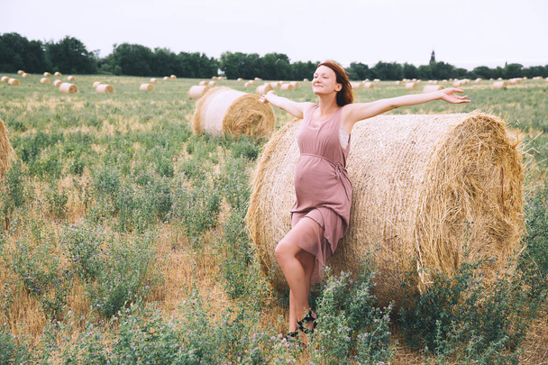Beautiful pregnant woman in dress on nature. Woman with raised arms on background of wheat field with haystacks at summer day. Photo of pregnancy, maternity, expectation. Mother waiting of a baby - Φωτογραφία, εικόνα
