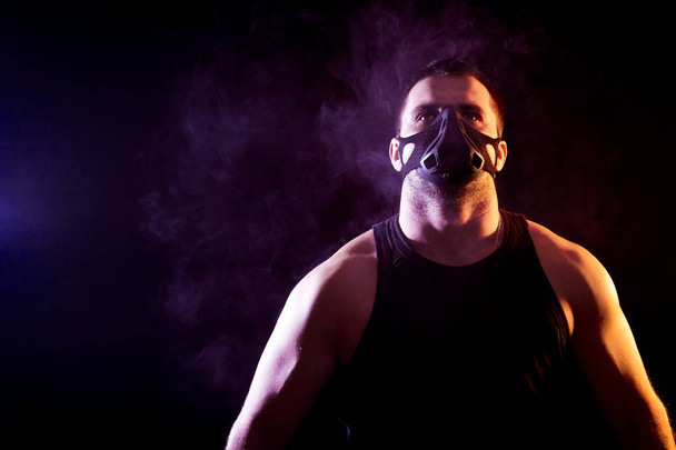 Ritratto di giovane atletico che indossa camicia sportiva e maschera da allenamento su sfondo nero isolato, intorno al fumo blu da un vaporizzatore
 - Foto, immagini