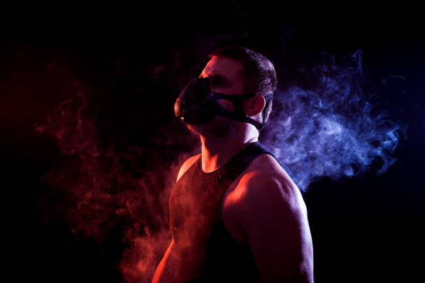 A young athletic man in a sports shirt breathes through a training black mask around a blue and red smoke cloud from a vape on a black isolated background - Zdjęcie, obraz