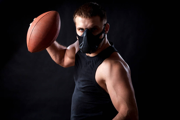 Retrato de jovem atlético vestindo camisa esporte, máscara de treinamento e segurando bola de rugby no fundo isolado preto
 - Foto, Imagem