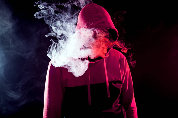The man in the hood smoke an electronic cigarette  on a background of white smoke - Foto, imagen