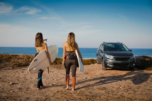 Two beautiful surfer girls near coastline with car getting ready for surfing - Фото, изображение