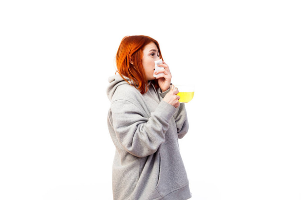 Μια νεαρή κοκκινομάλλα γυναίκα σε ένα γκρι φούτερ είναι άρρωστος, φτερνίζεται και ποτά από ένα μεγάλο διαφανές φλιτζάνι αντιϊική πράκτορα για θεραπεία σε λευκό φόντο απομονωμένες - Φωτογραφία, εικόνα