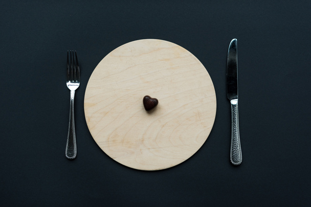 top view of heart shaped chocolate candy on round board with cutlery isolated on black - Zdjęcie, obraz