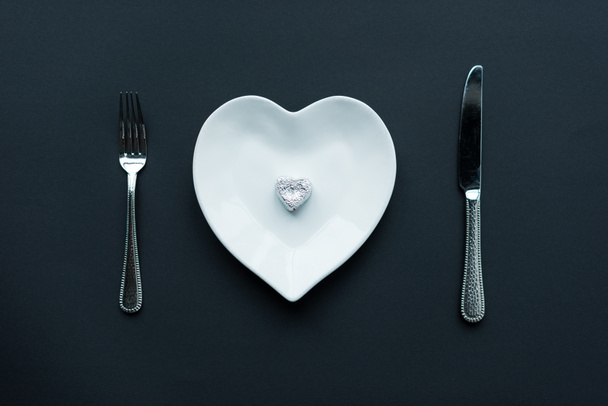 κάτοψη του τυλιγμένη καραμέλα σε λευκό σχήμα καρδιάς πλάκα με τα μαχαιροπήρουνα που απομονώνονται σε μαύρο - Φωτογραφία, εικόνα