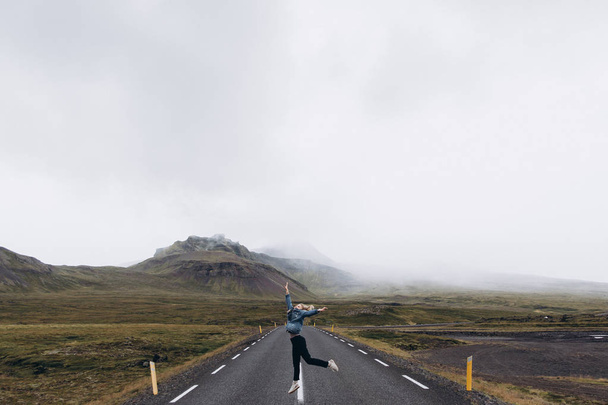 Vue latérale de la jeune femme sautant sur la route par temps froid et pluvieux sur fond de montagnes
 - Photo, image