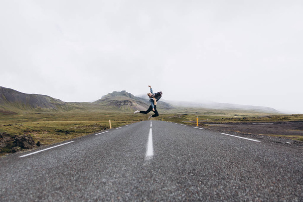 Side view of young woman jumping on the road in cold rainy day on mountains background - Fotó, kép