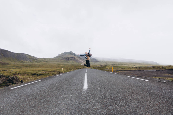 Vue latérale de la jeune femme sautant sur la route par temps froid et pluvieux sur fond de montagnes
 - Photo, image