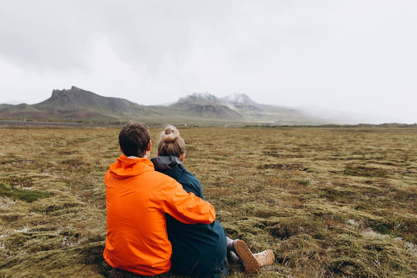 Nuori pari istuu niityllä kylmässä sateinen päivä ja nauttia vuoristomaisema
 - Valokuva, kuva