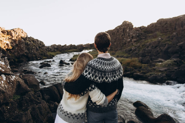 Νεαρό ζευγάρι ντυμένα σε ζεστό πλεκτό πουλόβερ αγκάλιασμα και ψάχνει για ποταμάκι - Φωτογραφία, εικόνα
