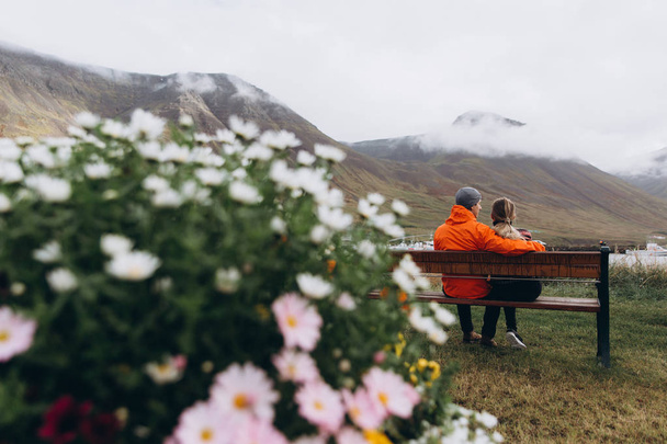 Вид сзади на молодую пару, сидящую на скамейке и наслаждающуюся моментом и цветами на переднем плане
 - Фото, изображение