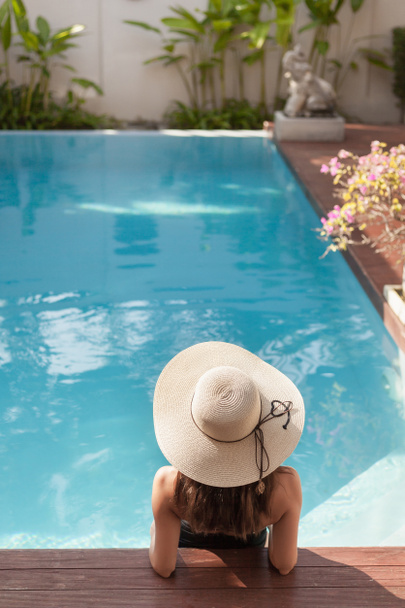 Achteraanzicht van de jonge vrouw in badpak en hoed zitten in het zwembad van hotel - Foto, afbeelding