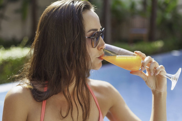 beautiful young woman in bikini and sunglasses drinking cocktail at poolside - Фото, зображення