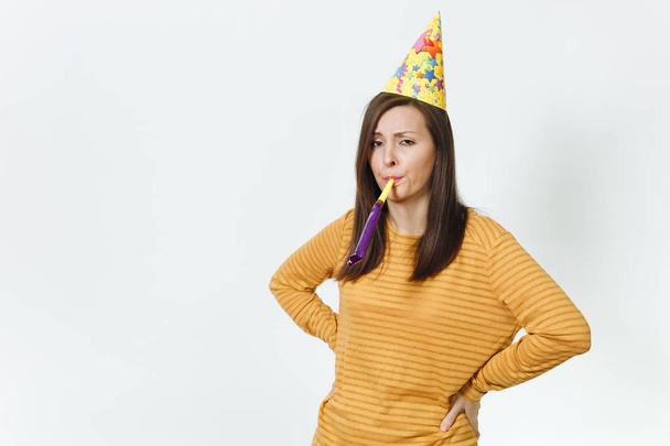 Красивая кавказская грустная молодая женщина в жёлтой одежде и шляпе на день рождения с каштановыми длинными волосами, играющая на трубе, празднующая праздник в одиночестве без подарка на белом фоне изолированная для рекламы
. - Фото, изображение