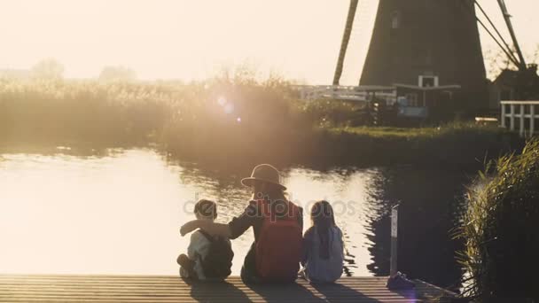 Woman with two kids sit on a lake pier on sunset. Amazing shot of a family together near water edge. 4K back view. - Кадры, видео