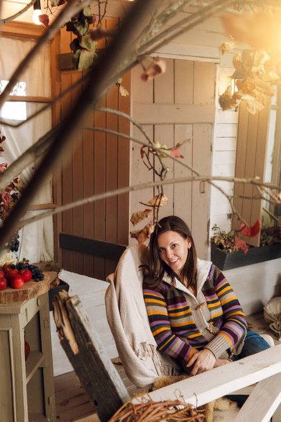 Beautiful young happy brown-haired woman, dressed in a sweater, jeans and yellow shoes, sitting on the porch in chair in autumn time. Courtyard, lit by flashlights, with dry fall leaves and pumpkins. - Foto, Imagen