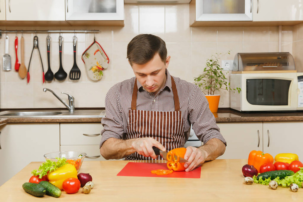 Knappe lachende Kaukasische jongeman in een schort, bruin shirt zitten aan tafel, snijdt groente voor salade met mes in lichte keuken. Dieet concept. Gezonde levensstijl. Thuis koken. Eten bereiden. - Foto, afbeelding