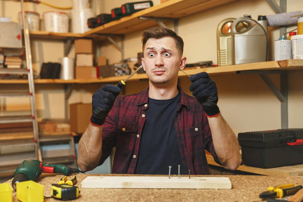 Αρκετά διασκεδαστικό ξεμωραμένος zonked Καυκάσιος νεαρός άνδρας στο καρό πουκάμισο, μαύρο T-shirt, γάντια συστροφή από κατσαβίδι βίδα, εργάζεται στο ξυλουργείο τόπο ξύλινο τραπέζι με κομμάτι του ξύλου, εργαλεία. - Φωτογραφία, εικόνα