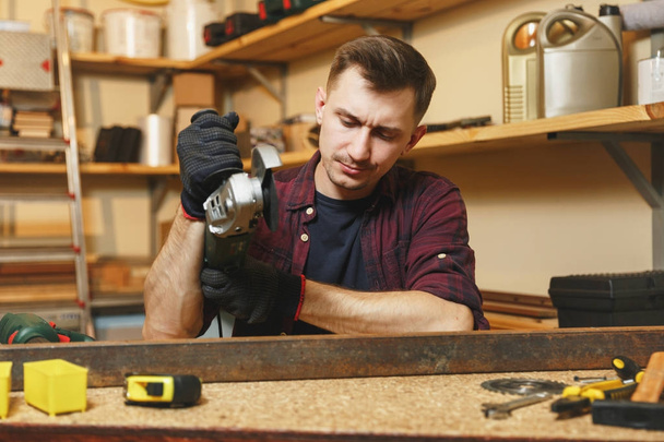 Handsome smiling caucasian young man in plaid shirt, black T-shirt, noise insulated headphones working in carpentry workshop at wooden table place with different tools, sawing iron with power saw. - Photo, Image