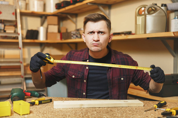 Handsome caucasian young man in plaid shirt, black T-shirt, gloves measuring length of piece of wood with tape measure, working in carpentry workshop at wooden table place with different work tools. - Photo, Image