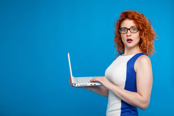 Isolated portrait of a business girl holding a laptop. on a blue background. The girl looks surprised and confused. Place for text. Business and emotional concept - Φωτογραφία, εικόνα