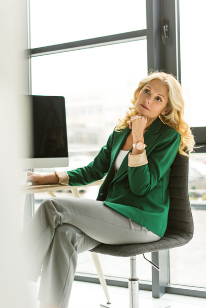 selective focus of pensive middle aged businesswoman sitting and looking away at workplace - Fotoğraf, Görsel
