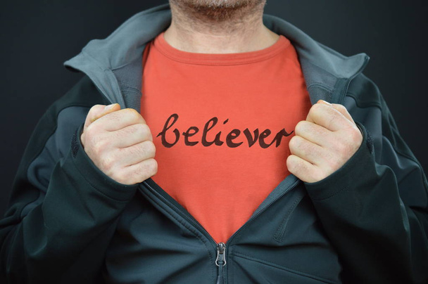 een man toont het woord gelovige geschreven op zijn rode t-shirt onder zijn zwarte jas - Foto, afbeelding