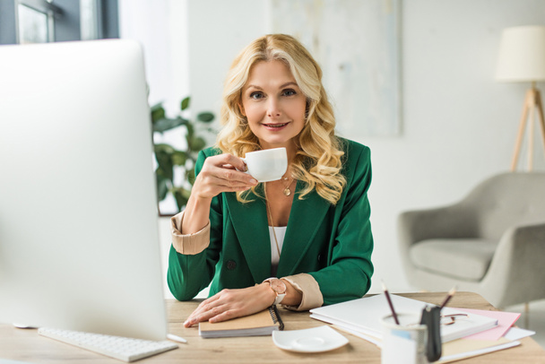 smiling middle aged businesswoman drinking coffee and using desktop computer at workplace - Photo, Image