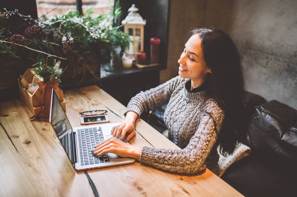 mooie jonge brunette vrouw met behulp van een laptop bij coffeeshop op een houten tafel in de buurt van venster tekst typen op een toetsenbord. In de winter, met het licht van de lamp, is ze gekleed in een warme grijze trui - Foto, afbeelding