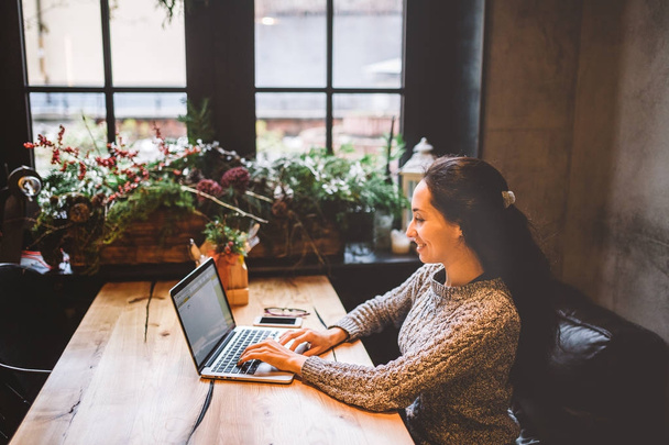 mooie jonge brunette vrouw met behulp van een laptop bij coffeeshop op een houten tafel in de buurt van venster tekst typen op een toetsenbord. In de winter, met het licht van de lamp, is ze gekleed in een warme grijze trui - Foto, afbeelding