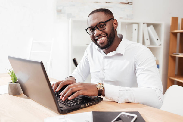 Retrato del sonriente hombre de negocios afroamericano en camisa blanca y gafas sentado y mirando alegremente en cámara mientras trabajaba en su computadora portátil en la oficina aislado
 - Foto, Imagen