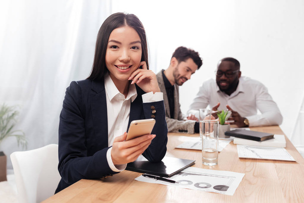 Portrait of beautiful asian businesswoman sitting in office and joyfully looking in camera with cellphone in hands and multinational business partners on background - Zdjęcie, obraz